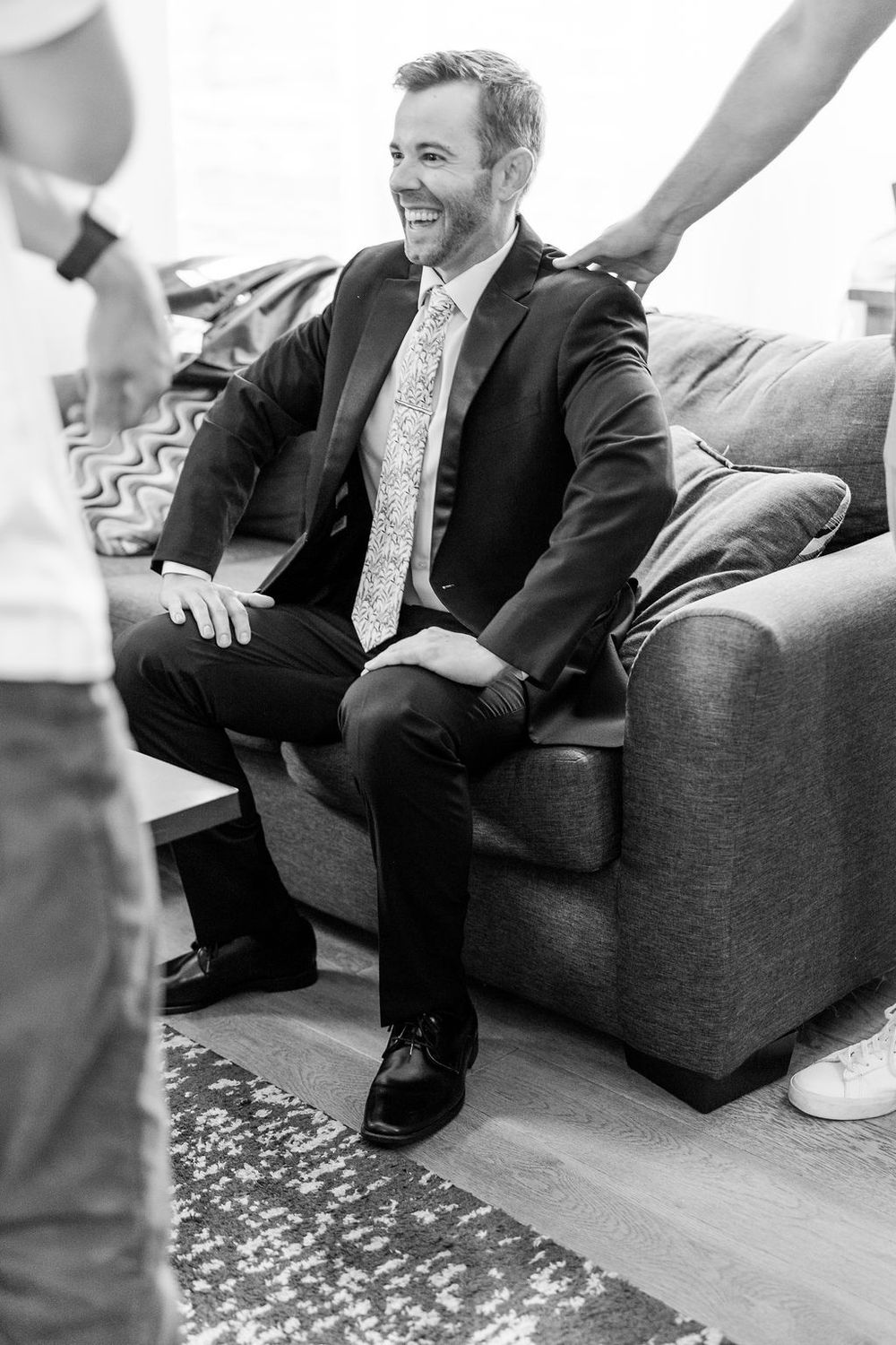 Wedding party members laugh while relaxing on gray couch during preparations.