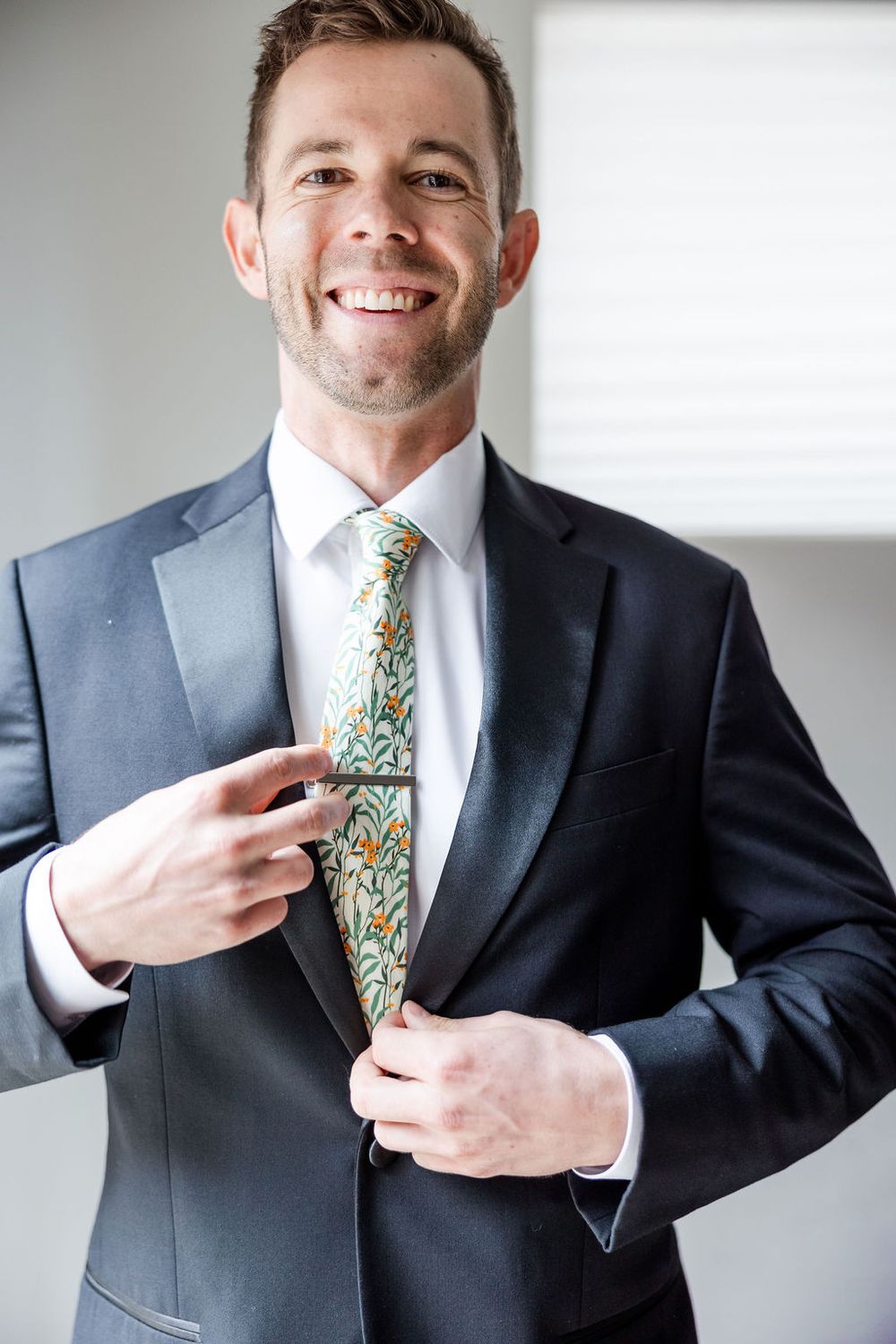 Professional in dark suit adjusts colorful floral patterned tie while standing against light background.