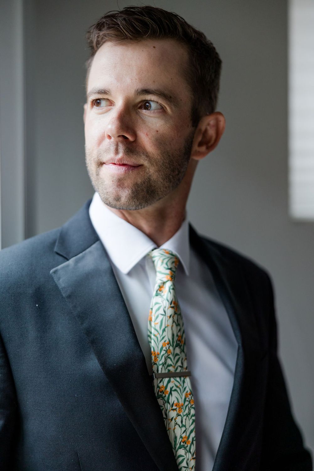 Professional portrait of person in navy suit with patterned tie against grey background.