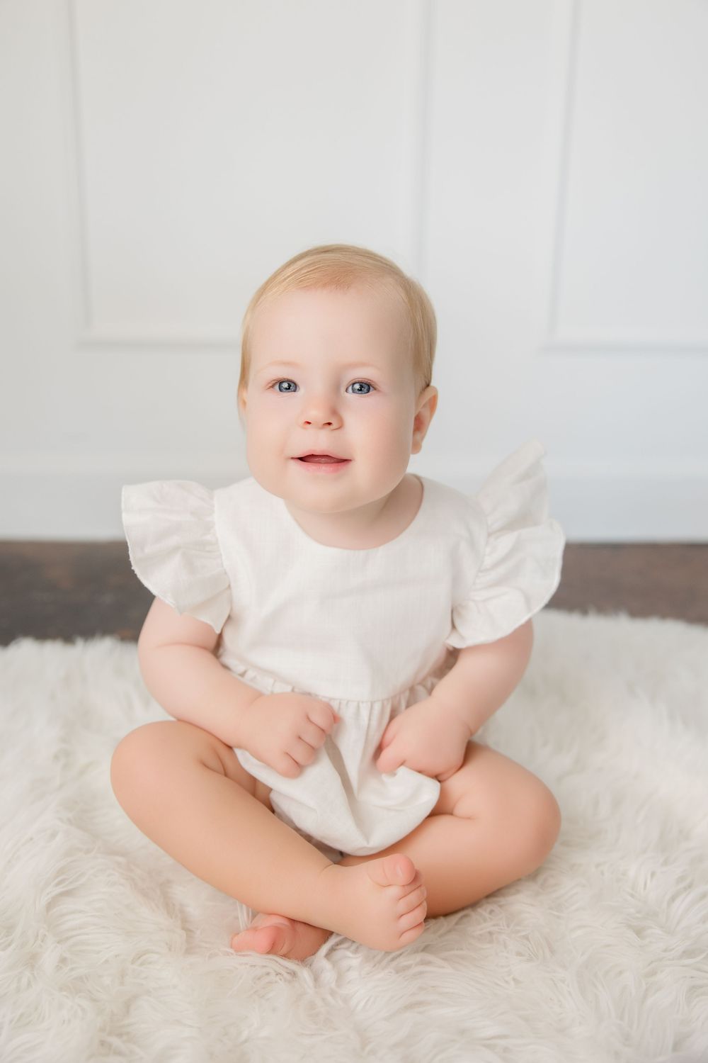 Sweet baby wearing white ruffled dress sitting on plush white rug during portrait session.