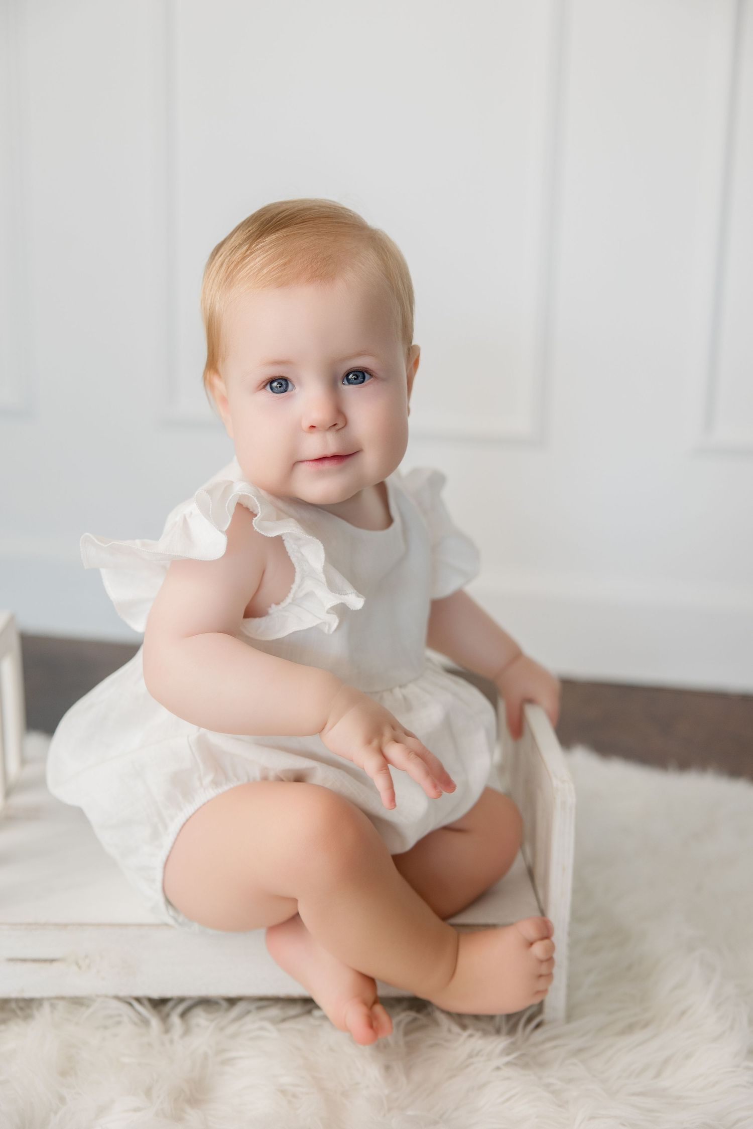 A baby in a white ruffled outfit sits on a soft white rug in three progressive studio photos.