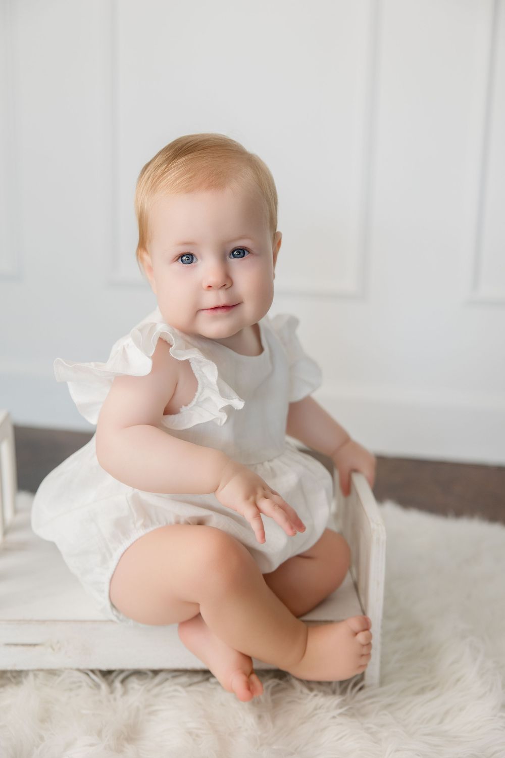 A baby in a white ruffled outfit sits on a soft white rug in three progressive studio photos.