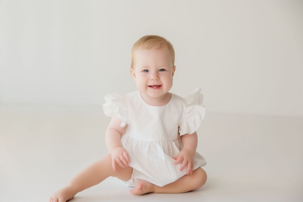 Baby in white ruffle dress sitting and smiling on light background during portrait session.
