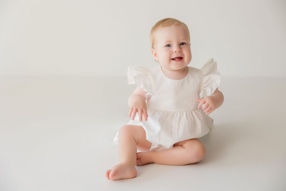 Sweet little baby in a white dress smiles while lying on a light gray backdrop during a portrait session.