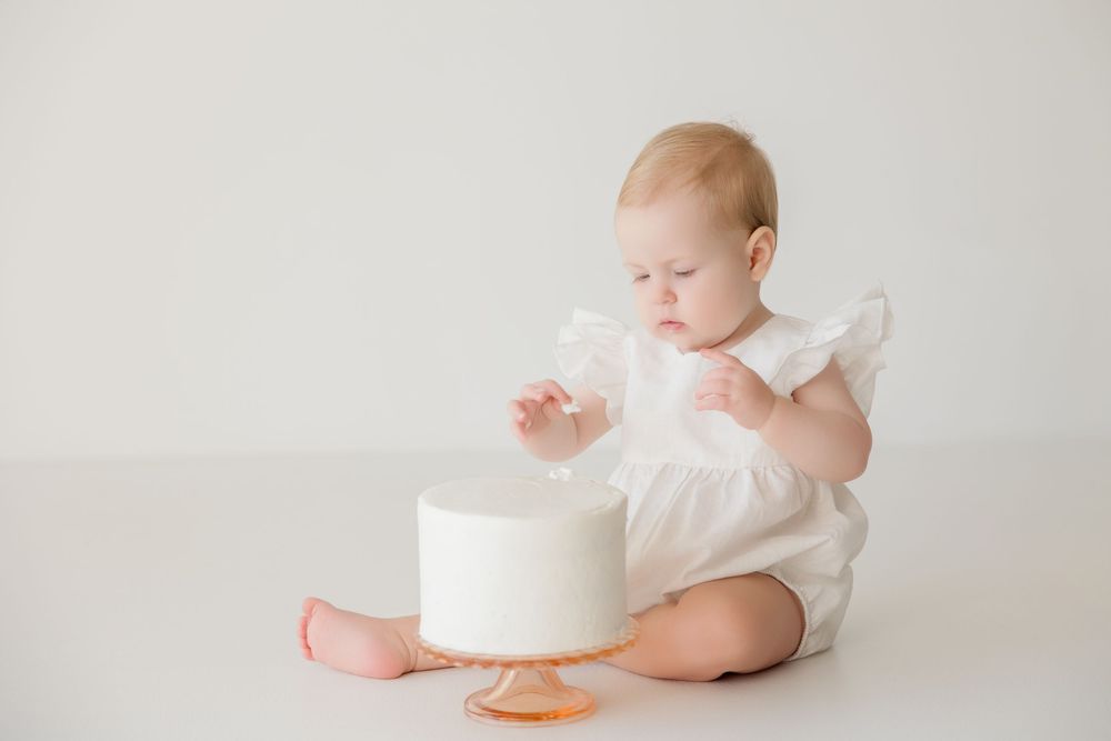 Baby in white outfit looking down at small white cake on light seamless backdrop.