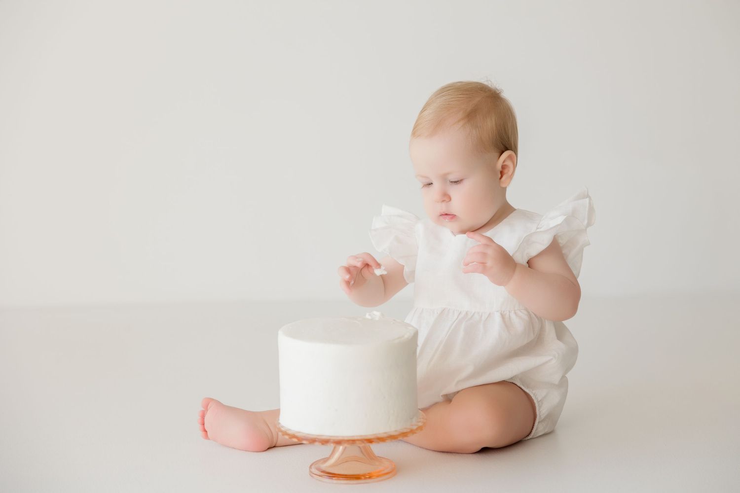 Baby in white outfit looking down at small white cake on light seamless backdrop.
