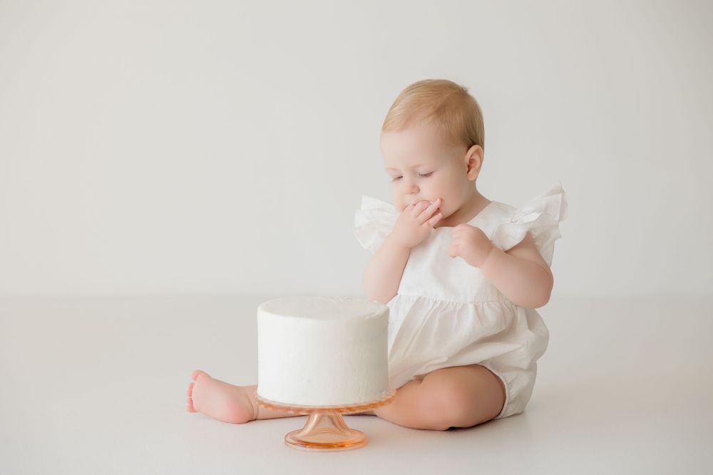 Baby in white outfit exploring cake and treats during cake smash photo session against neutral background.
