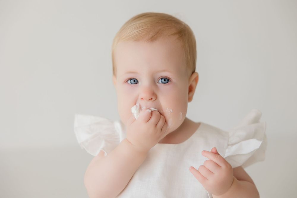 Baby in white dress with ruffled sleeves brings fingers to mouth while sitting against neutral background.