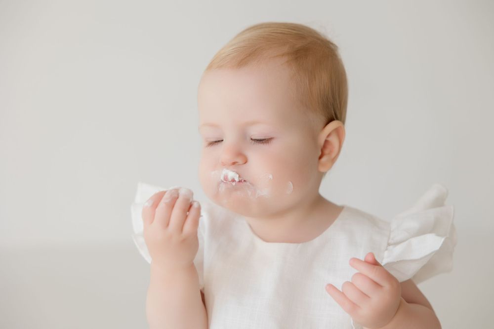 Baby in white dress smashes face into white cake during first birthday cake smash photo session.