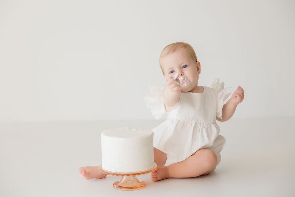 A baby in a white outfit lying on their side on a soft cream colored backdrop.