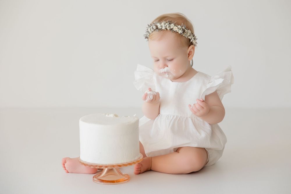 Baby wearing floral crown and white dress poses with white birthday cake on light backdrop.