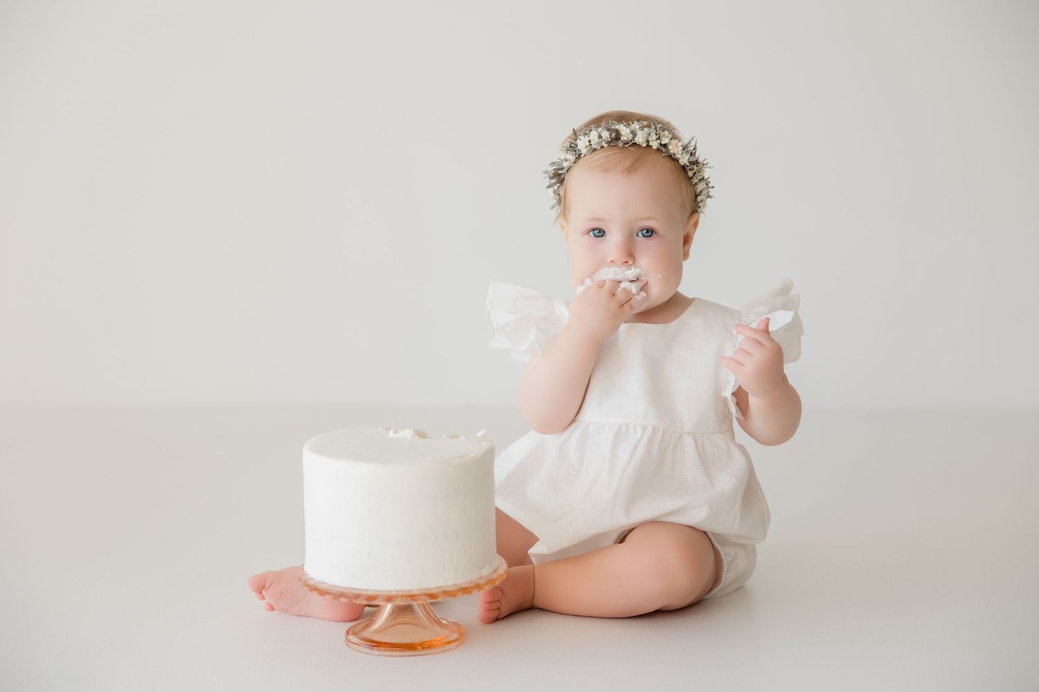Baby in white dress sitting with elegant white birthday cake on light backdrop in minimalist studio setup.