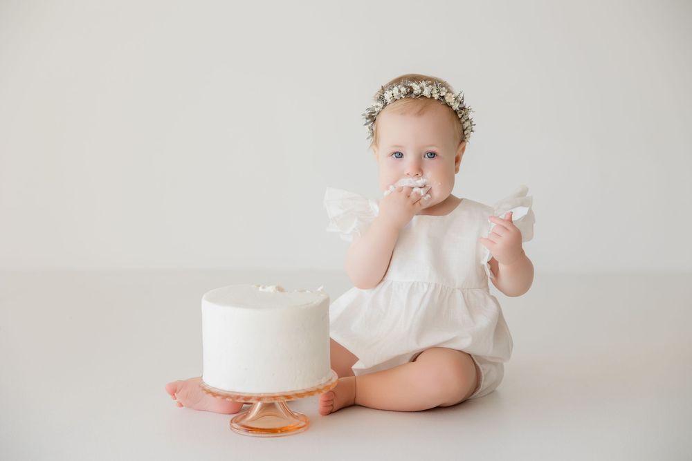 Baby in white dress sitting with elegant white birthday cake on light backdrop in minimalist studio setup.