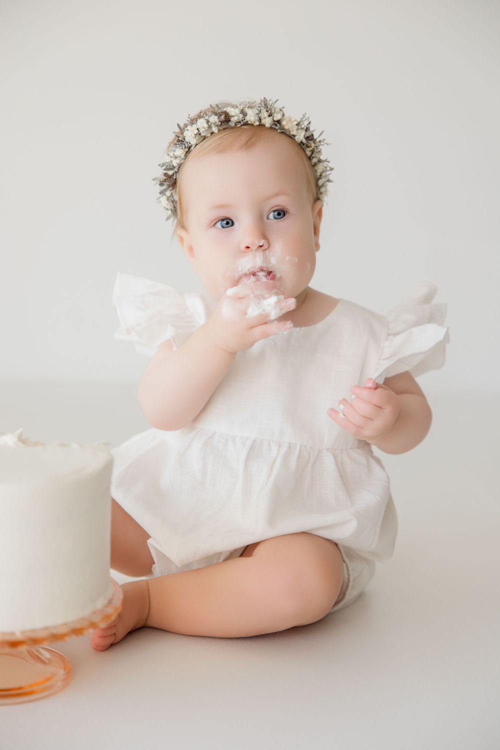 Baby wearing delicate floral crown and white outfit during celebratory photo session with cake.