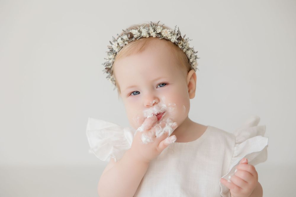 Baby in white outfit and floral crown enjoying cake during milestone photo session.