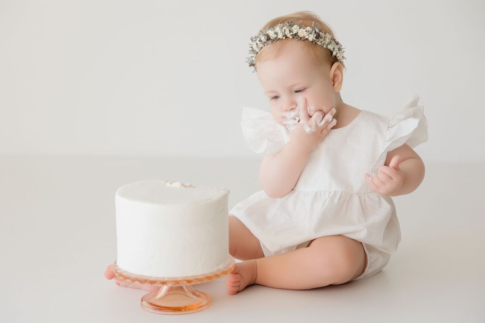 Baby in white outfit reaching towards simple white cake during minimalist milestone photo session.