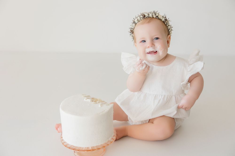 Baby in white ruffled dress smiles while sitting next to simple white cake.