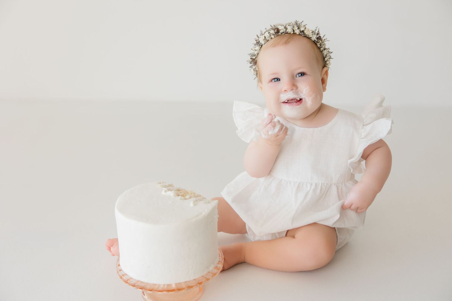 Baby in white ruffled dress smiles while sitting next to simple white cake.