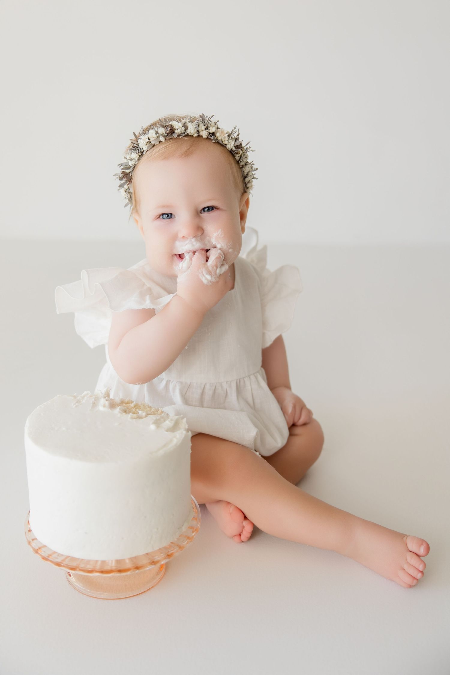 Baby in white dress explores white birthday cake during cake smash photography session.