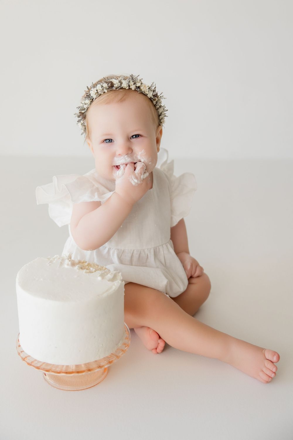 Baby in white dress explores white birthday cake during cake smash photography session.