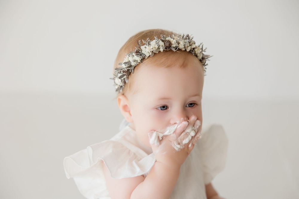 Minimalist portrait session of baby wearing white dress and floral crown against light backdrop.