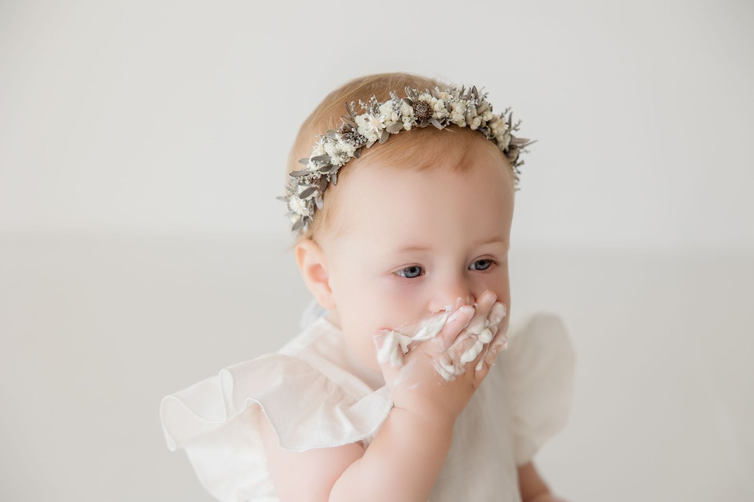 Minimalist portrait session of baby wearing white dress and floral crown against light backdrop.