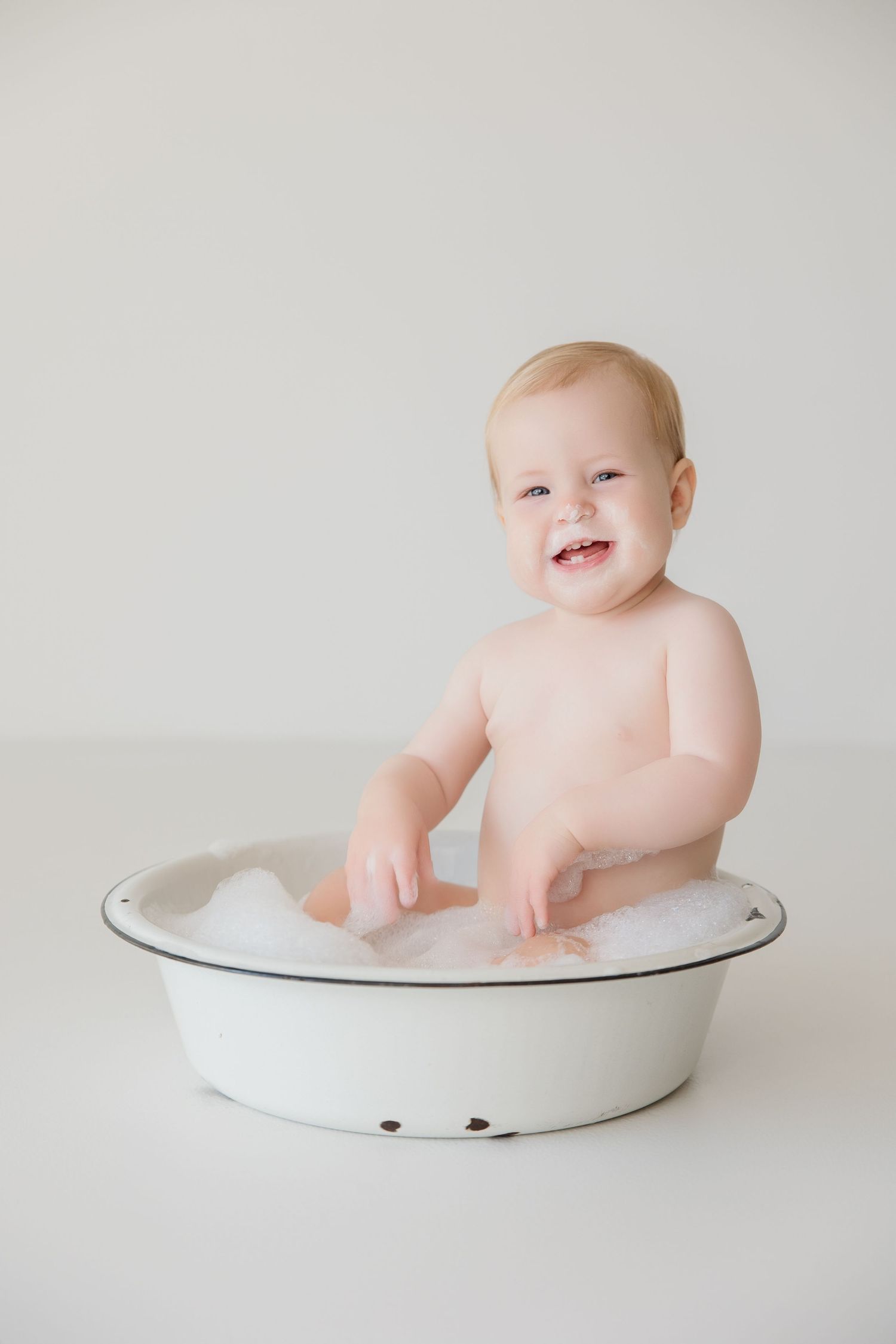 Smiling baby sitting in white enamel bowl during minimalist photo shoot.