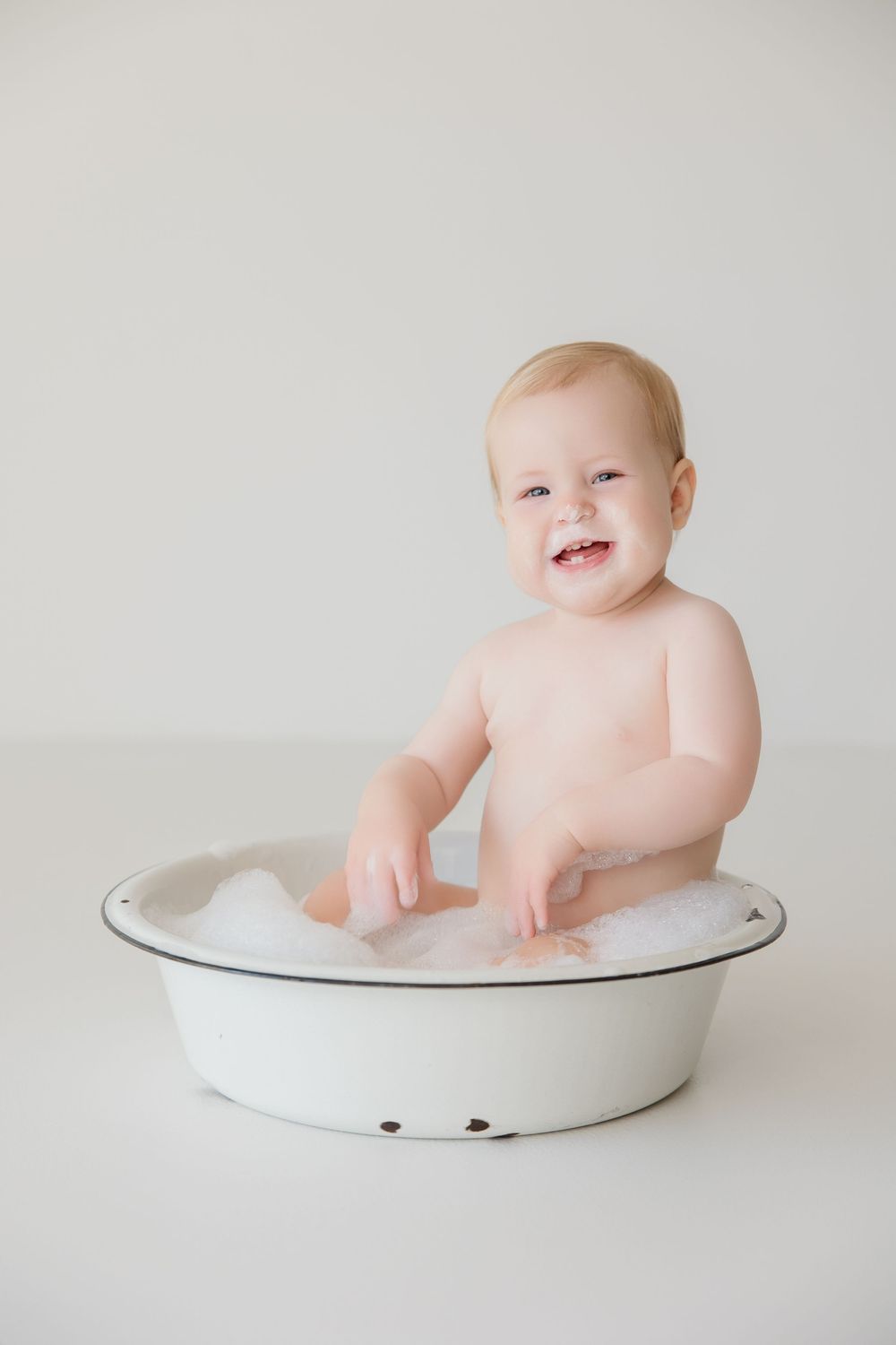 Smiling baby sitting in white enamel bowl during minimalist photo shoot.