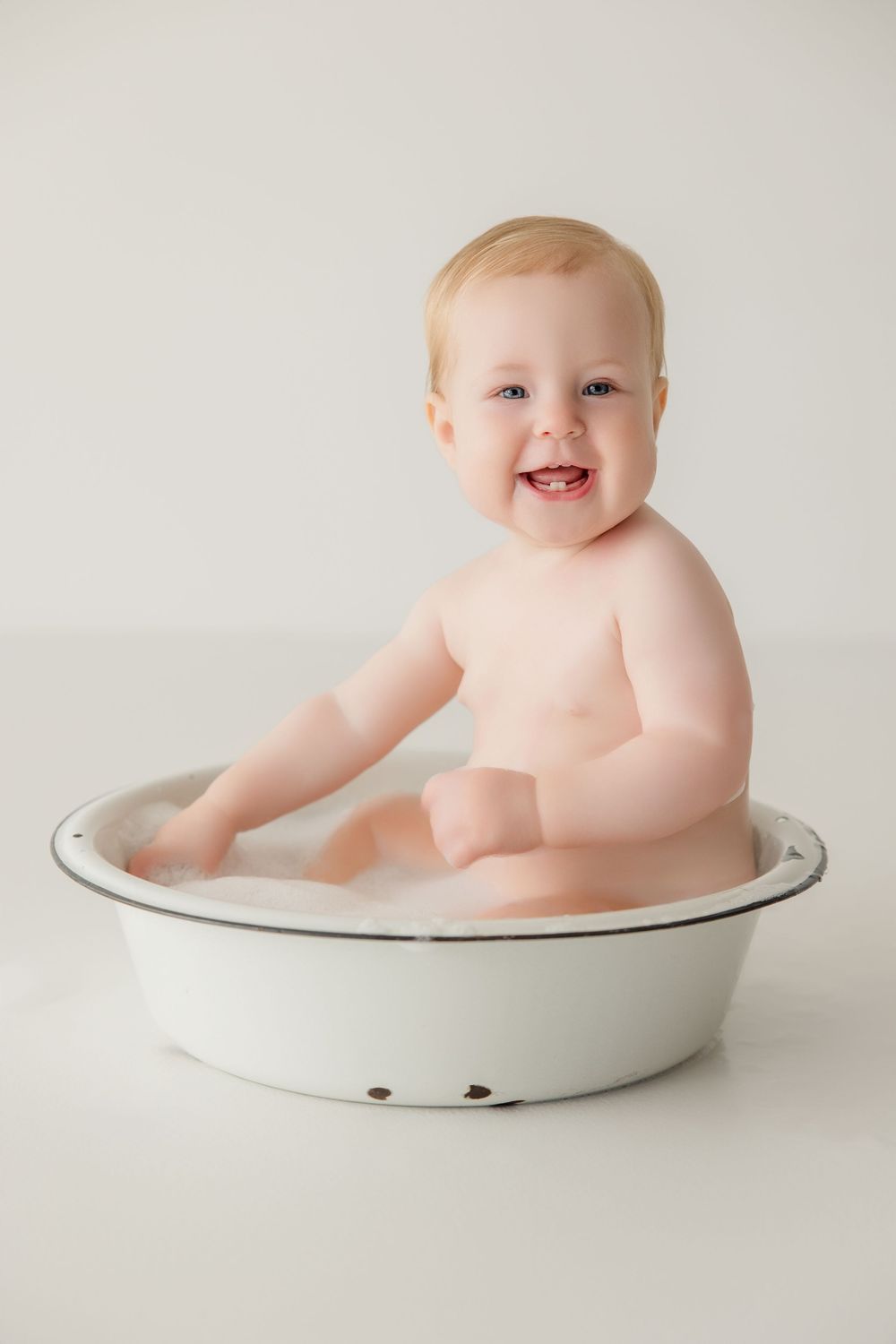 Two sweet milestone portraits show baby sitting in white enamel basin against neutral studio background.