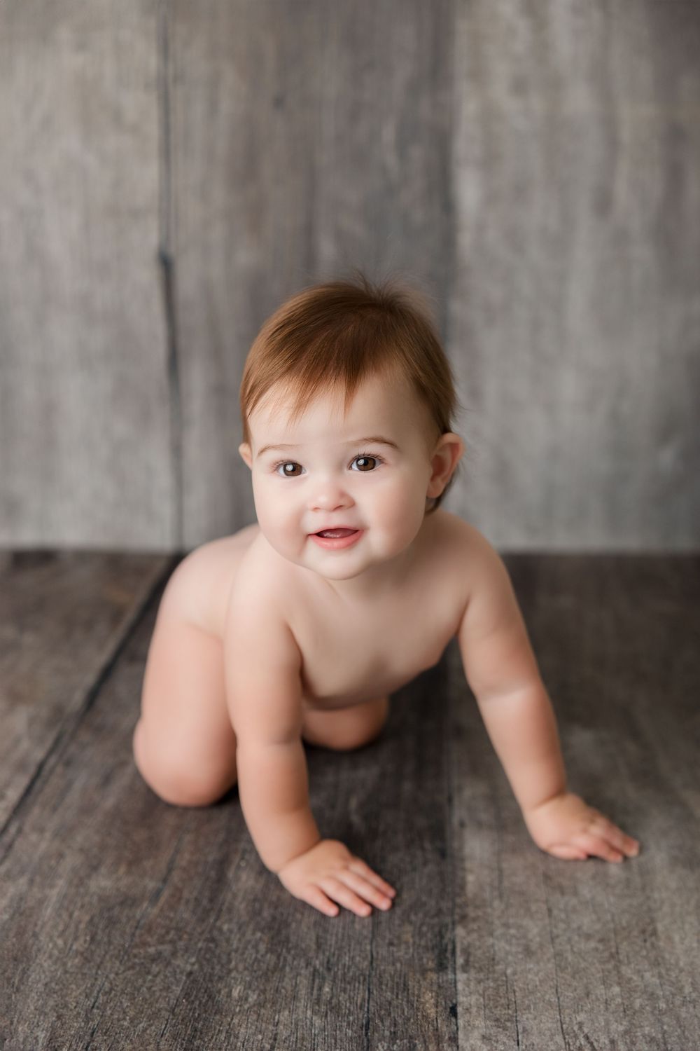 A baby wearing only a diaper crawls on a wooden floor against a gray wall background, smiling brightly.