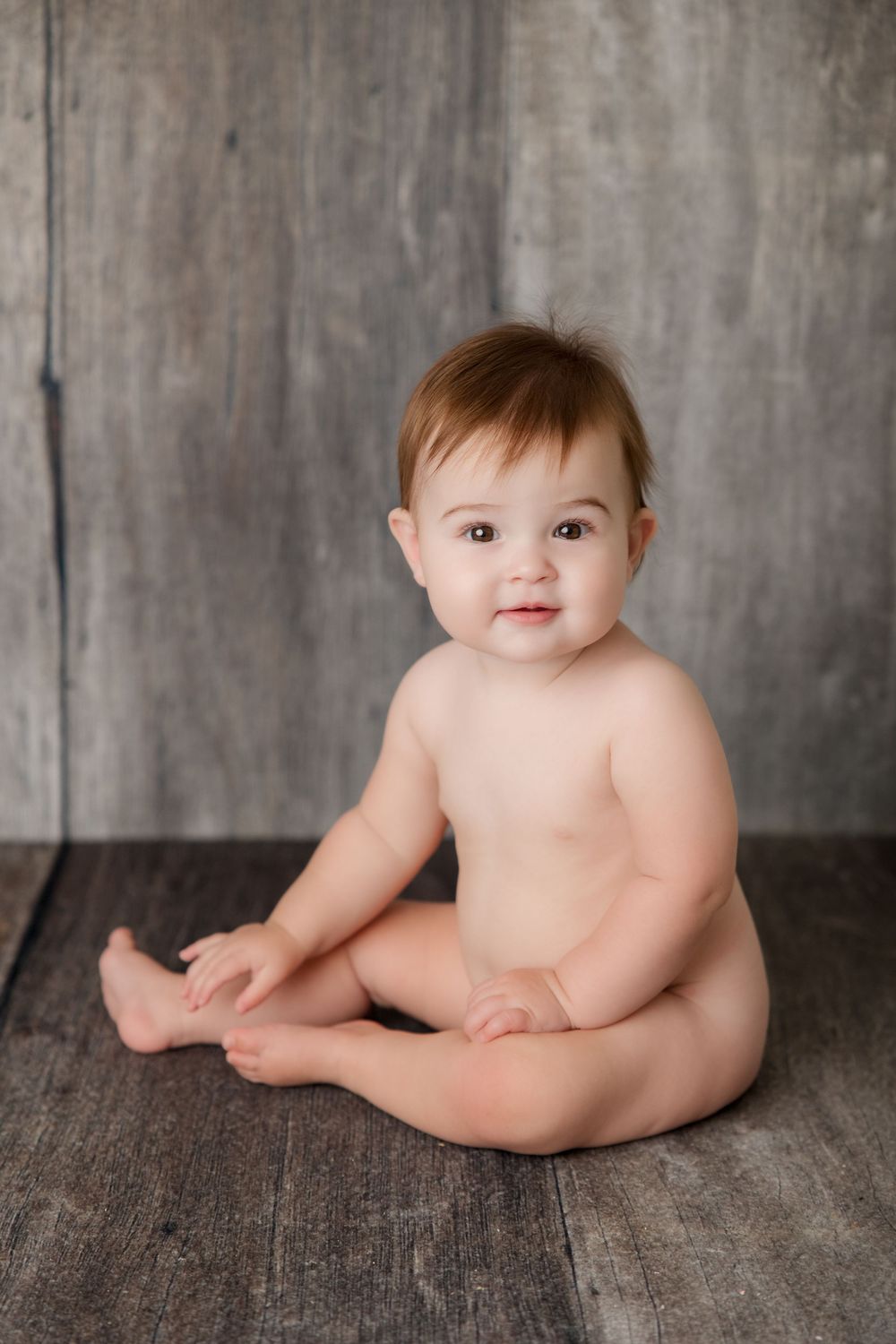 A chubby baby sits on wooden floor against grey backdrop during professional portrait session.