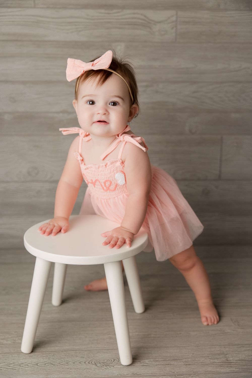 Small figure in peach tulle dress and bow stands at white stool against gray wooden backdrop.