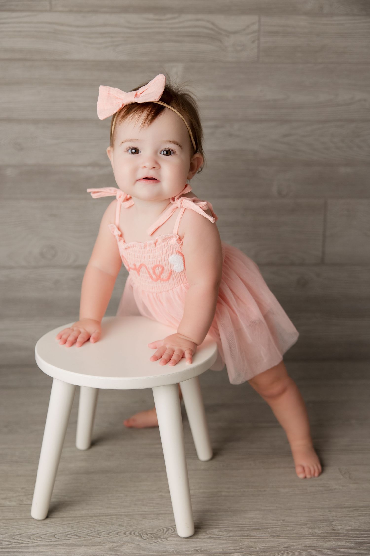 Small figure in peach tulle dress and bow stands at white stool against gray wooden backdrop.