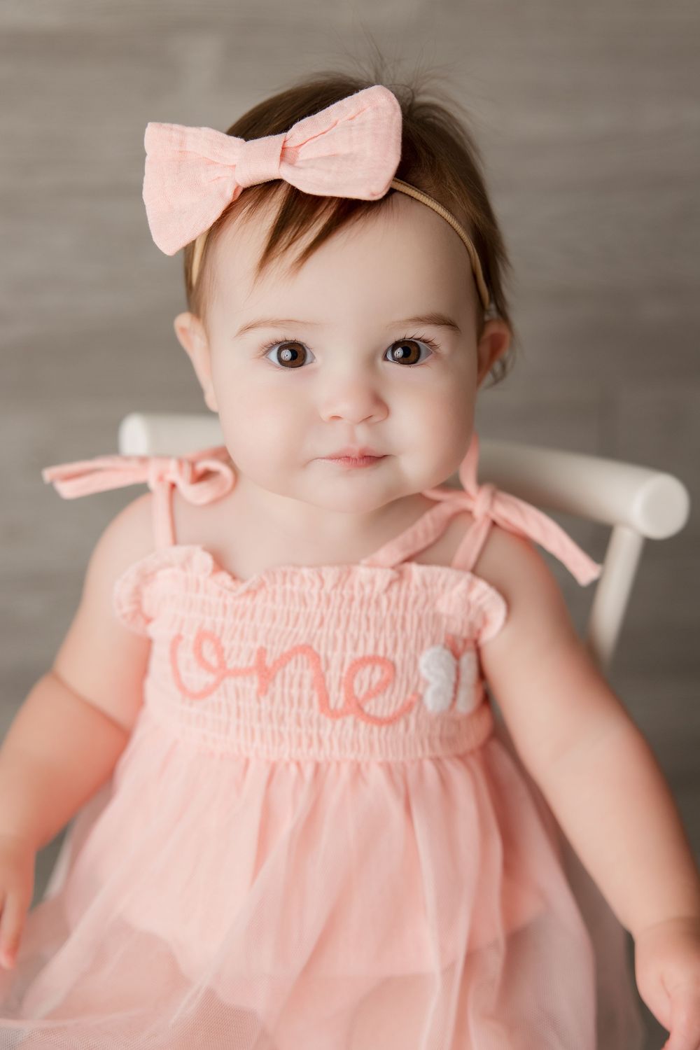 Baby wearing a delicate pink outfit with bow headband sits against a grey backdrop.