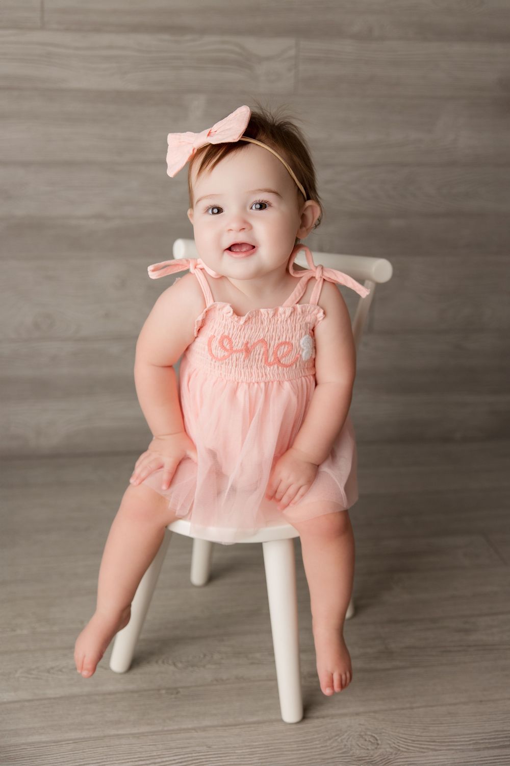 A baby in a peachy-pink dress sits on a white wooden chair against a gray backdrop in a photography session.
