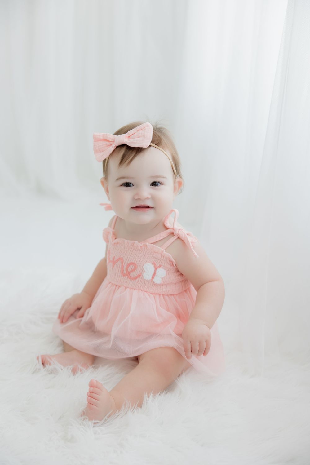 Baby in pink ruffle dress and bow laying on soft white blanket at professional photo studio session.