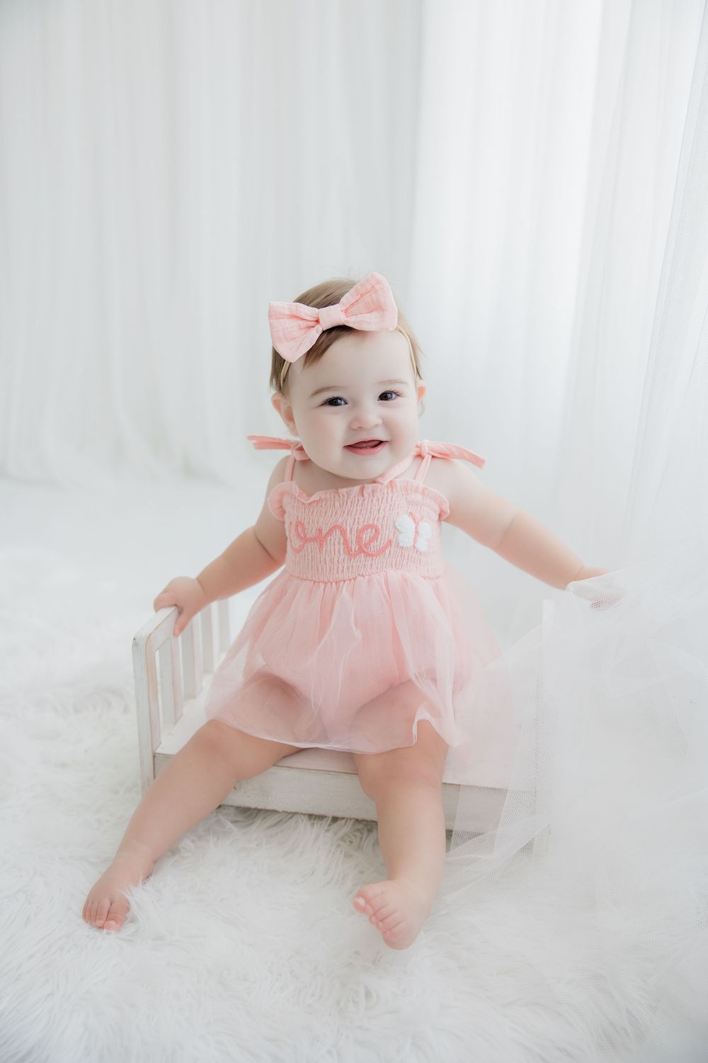 Baby in pink ruffled dress crawls and plays on soft white carpet during photography session.