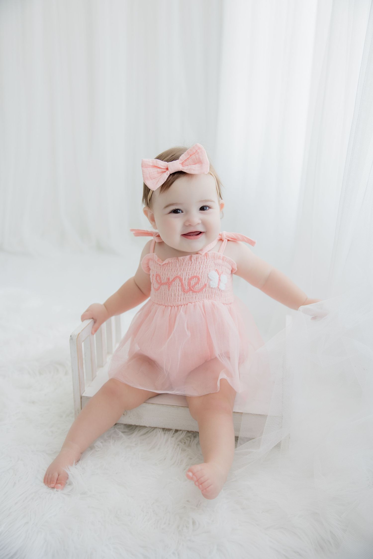 Baby in pink ruffled dress crawls and plays on soft white carpet during photography session.