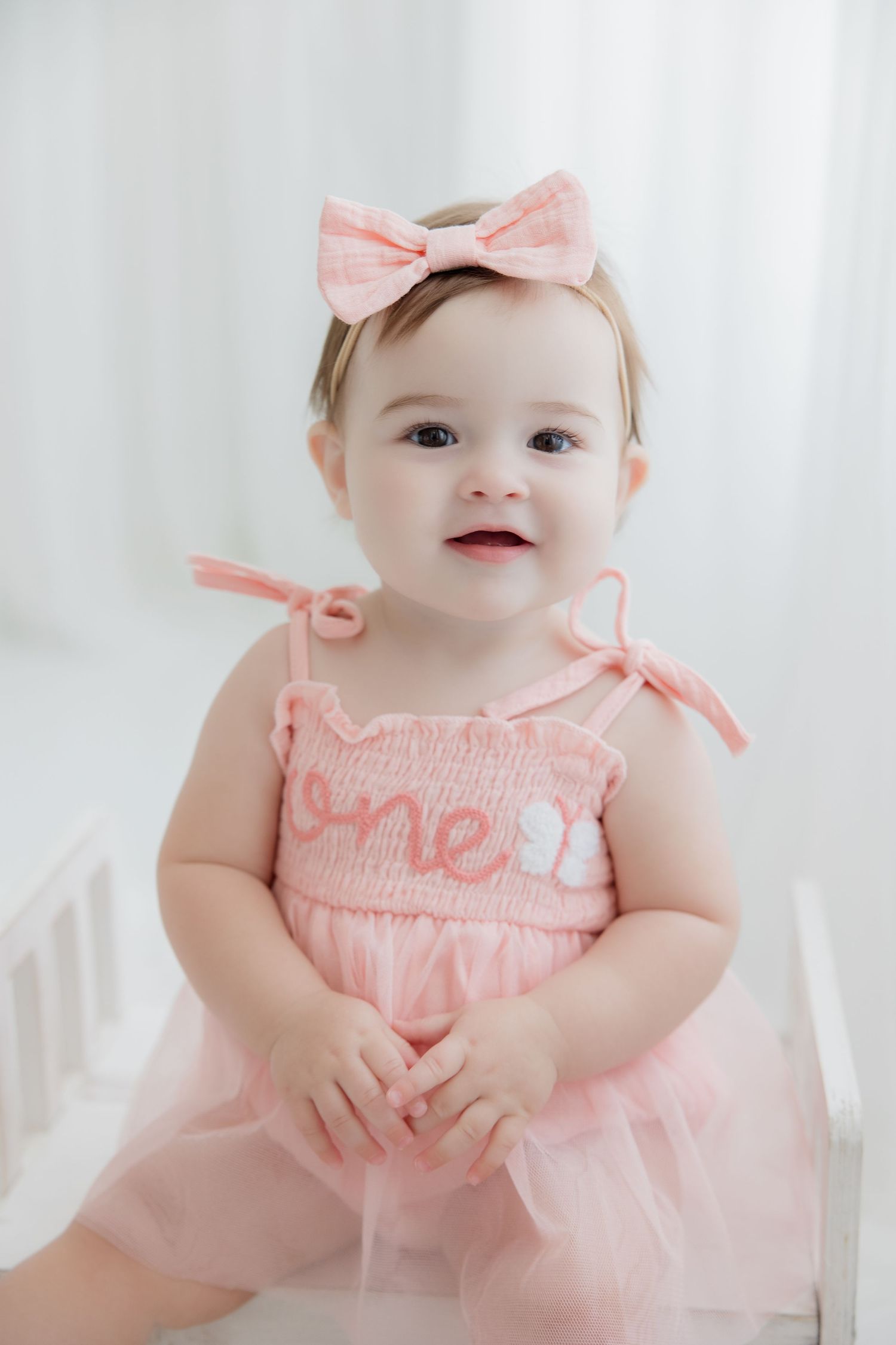 Baby wearing pink smocked dress with matching bow lounges on white backdrop in adorable portrait series.