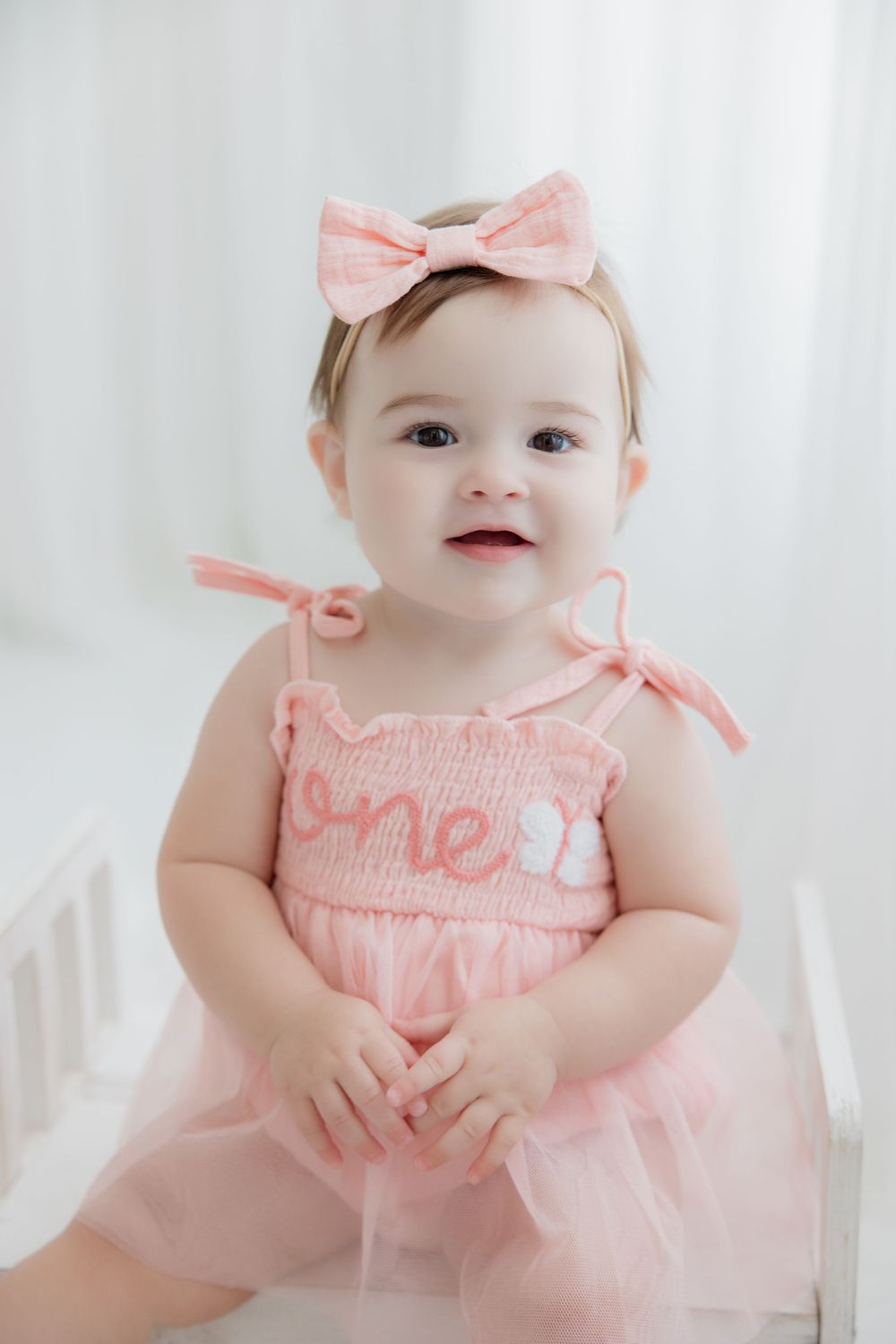Baby wearing pink smocked dress with matching bow lounges on white backdrop in adorable portrait series.