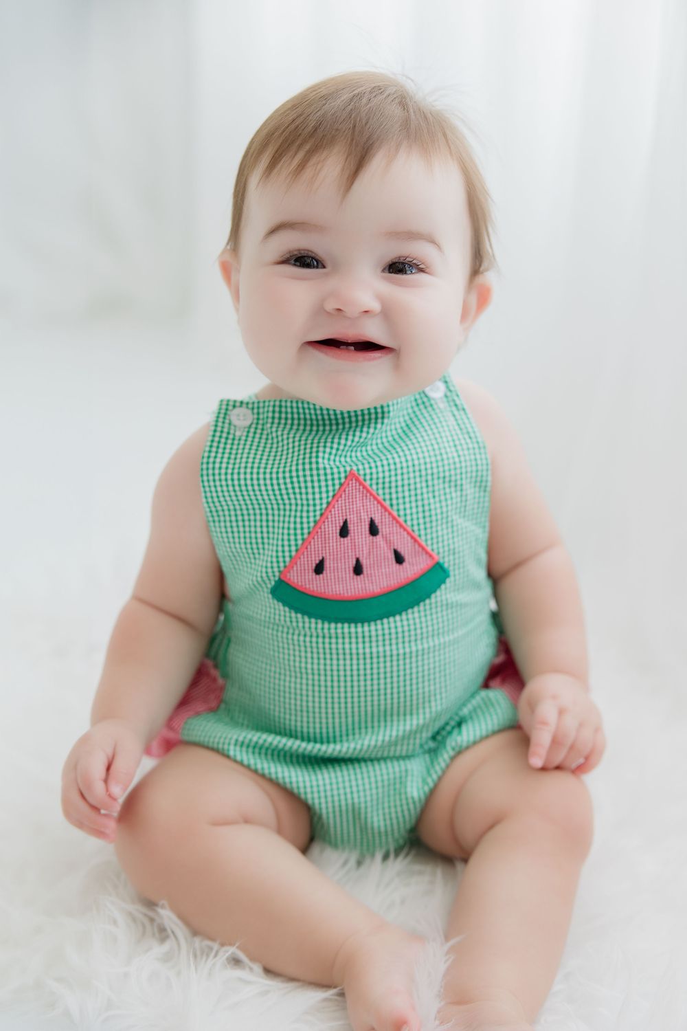 Infant in green gingham watermelon romper smiles while lying on white background.
