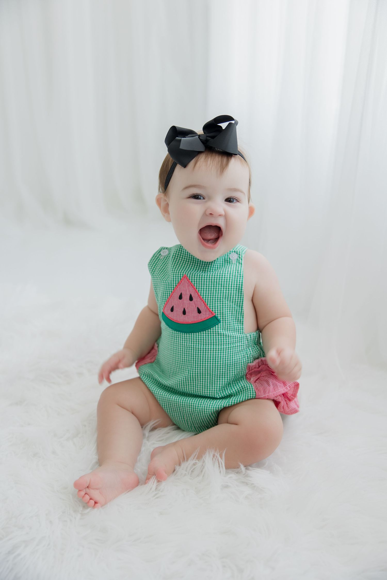 Baby wearing green watermelon romper poses on white fur rug in multiple candid shots showing joyful expressions.