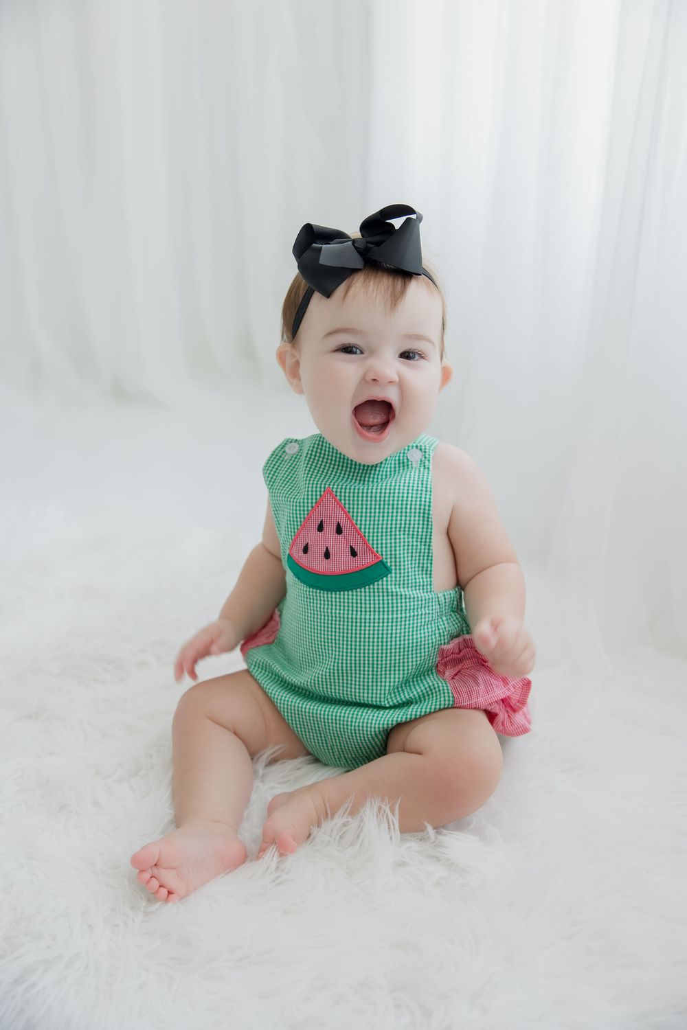 Baby wearing green watermelon romper poses on white fur rug in multiple candid shots showing joyful expressions.