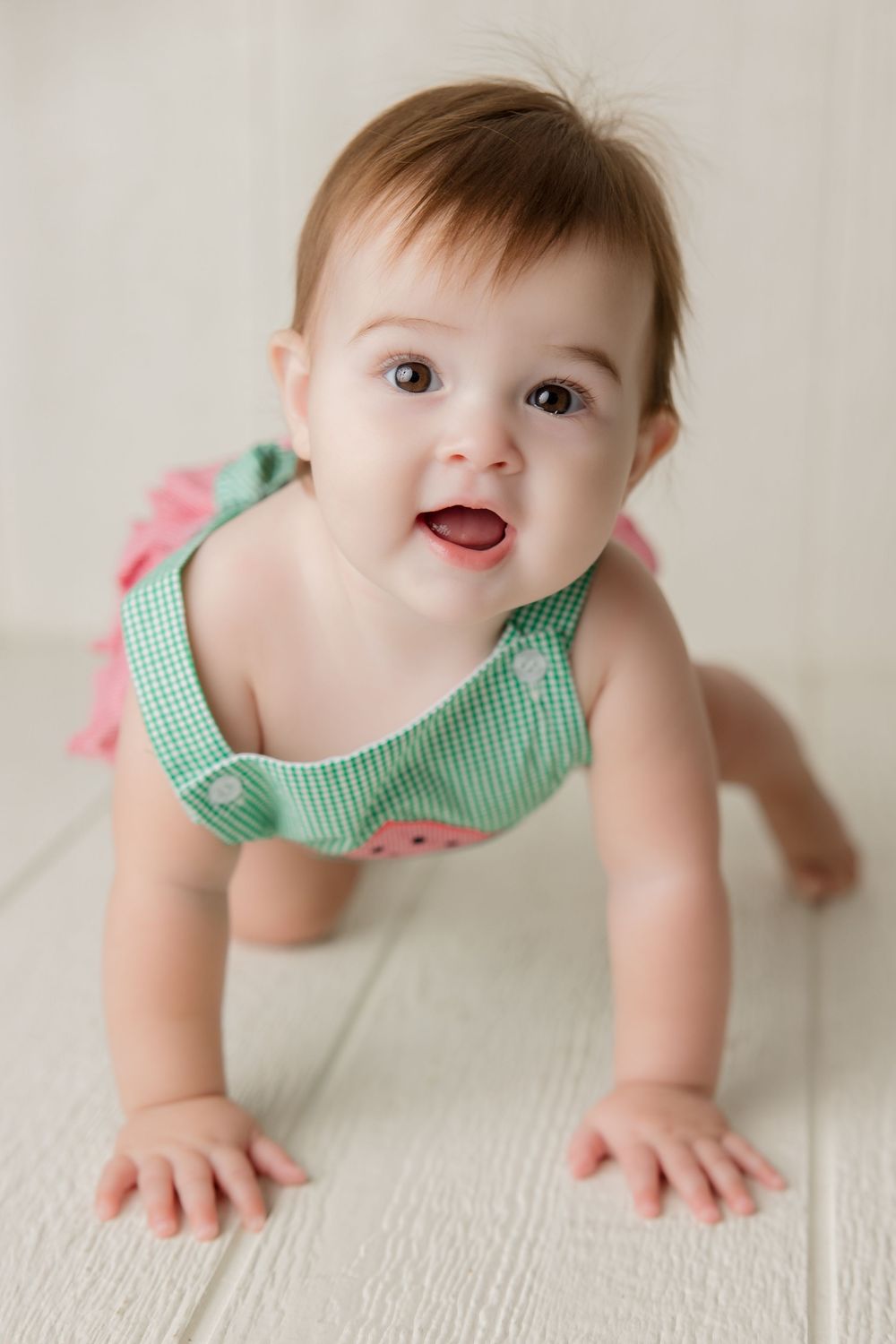 Baby wearing green watermelon outfit crawls playfully on white wooden floor.