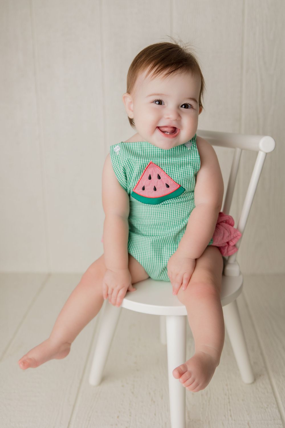 Infant in green gingham watermelon outfit poses on small white chair during photo session.