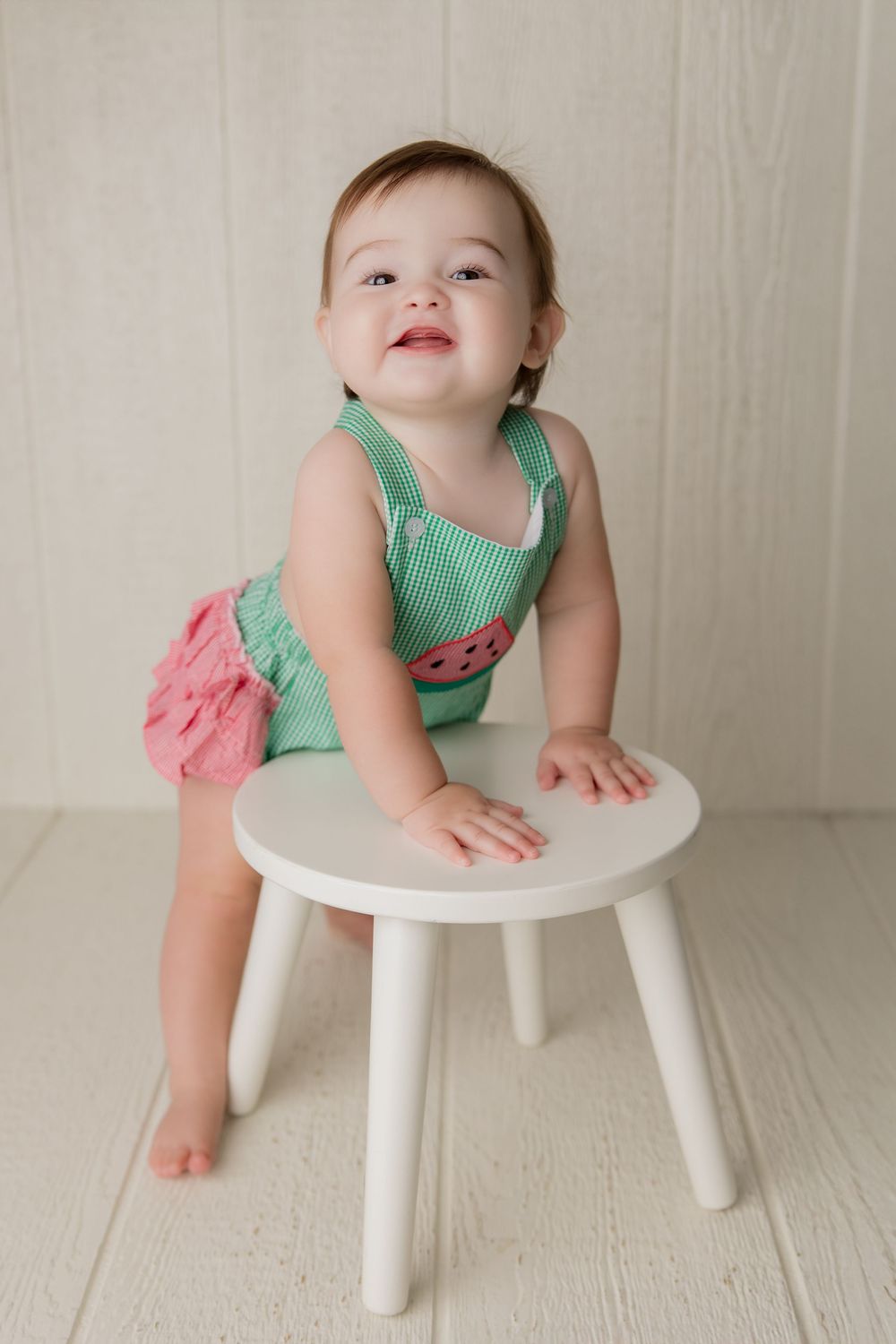 A smiling baby in a mint green outfit leans playfully over a small white wooden stool in a bright studio setting.