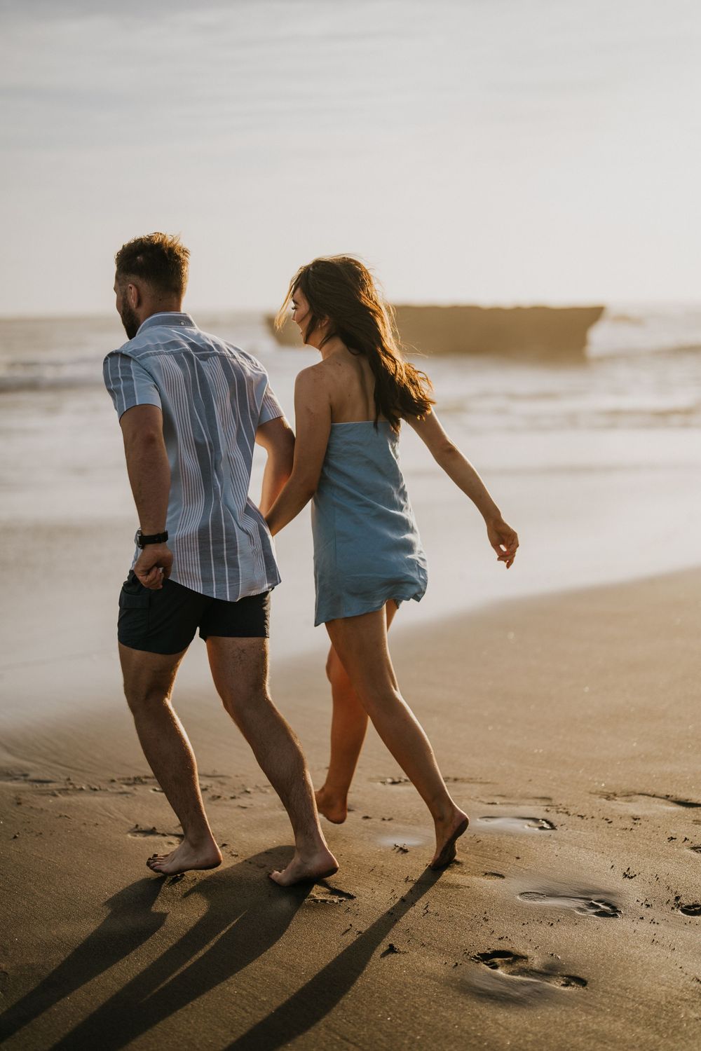 Two people run playfully along a sandy beach at golden hour casting long shadows.