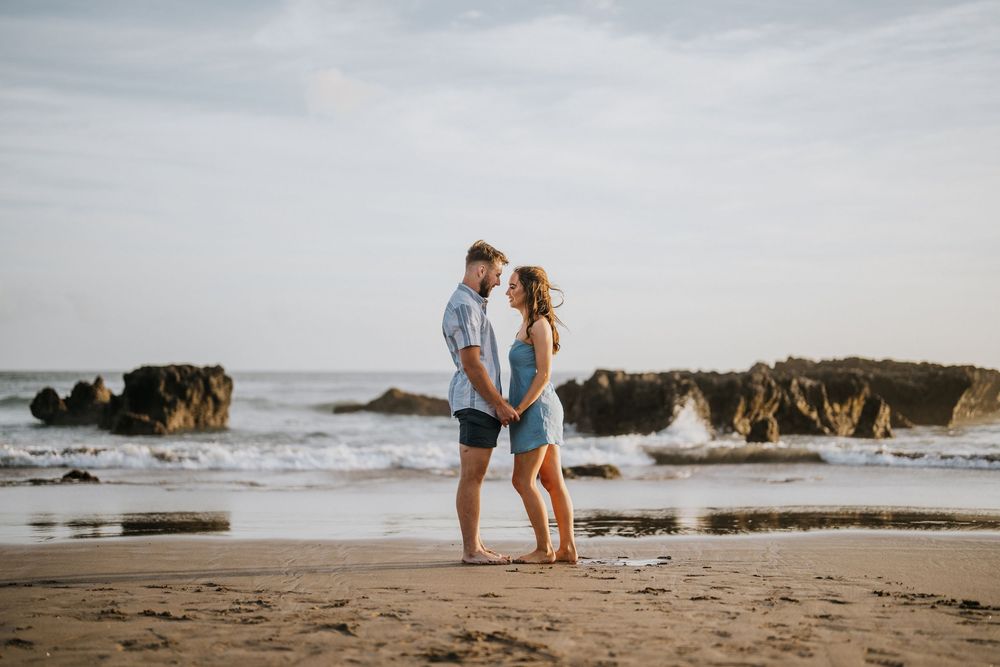 A couple walks hand in hand along a scenic beach with rocky cliffs in the distance.