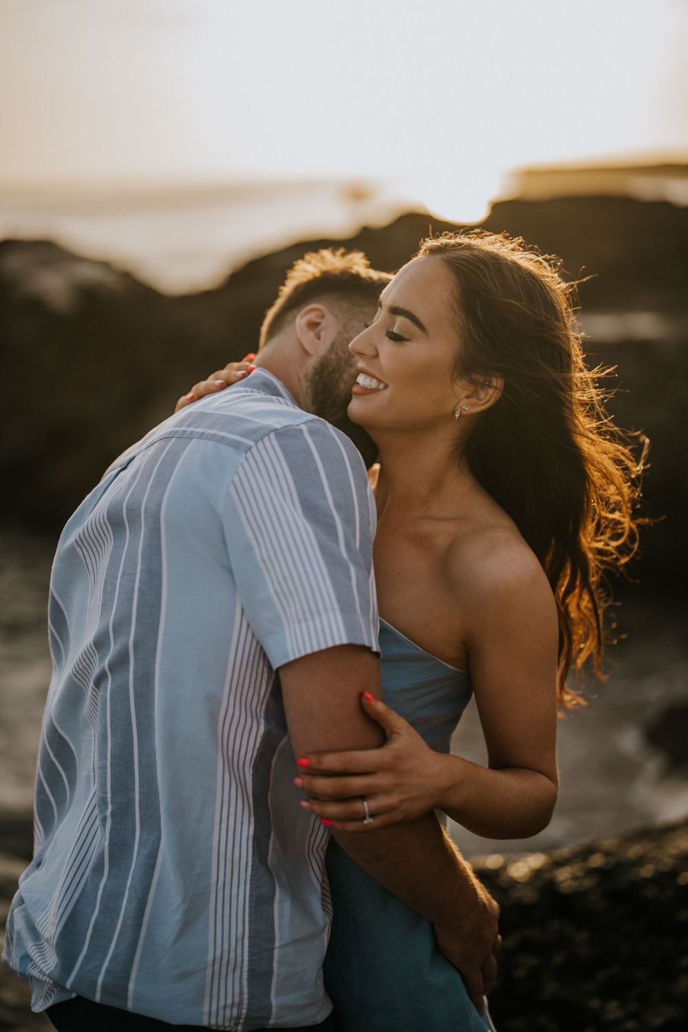 Two people share a tender embrace on the beach during golden hour with sun flares in the background.