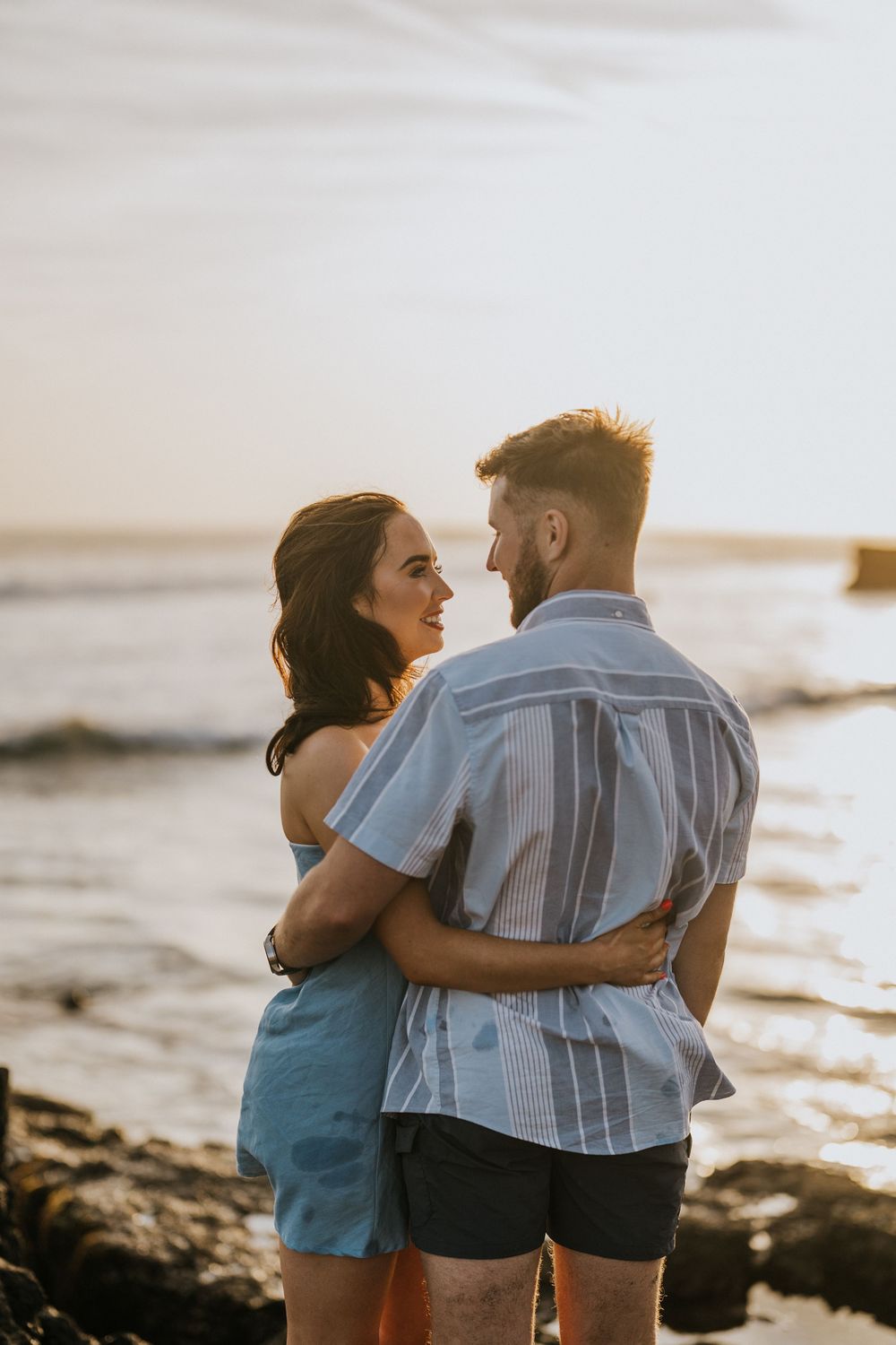 A couple embraces while standing on rocky coastal terrain at sunset.